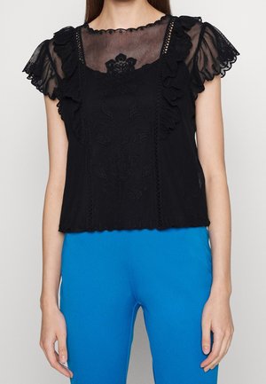 Femme portant un haut court en dentelle noire à volants avec broderie florale et un pantalon bleu vif sur un fond uni.