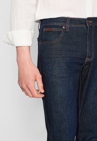 Mörkblå denimjeans med kontrastsömmar, en enda framficka och en läderpatch på midjebandet; hand vilande på låret.