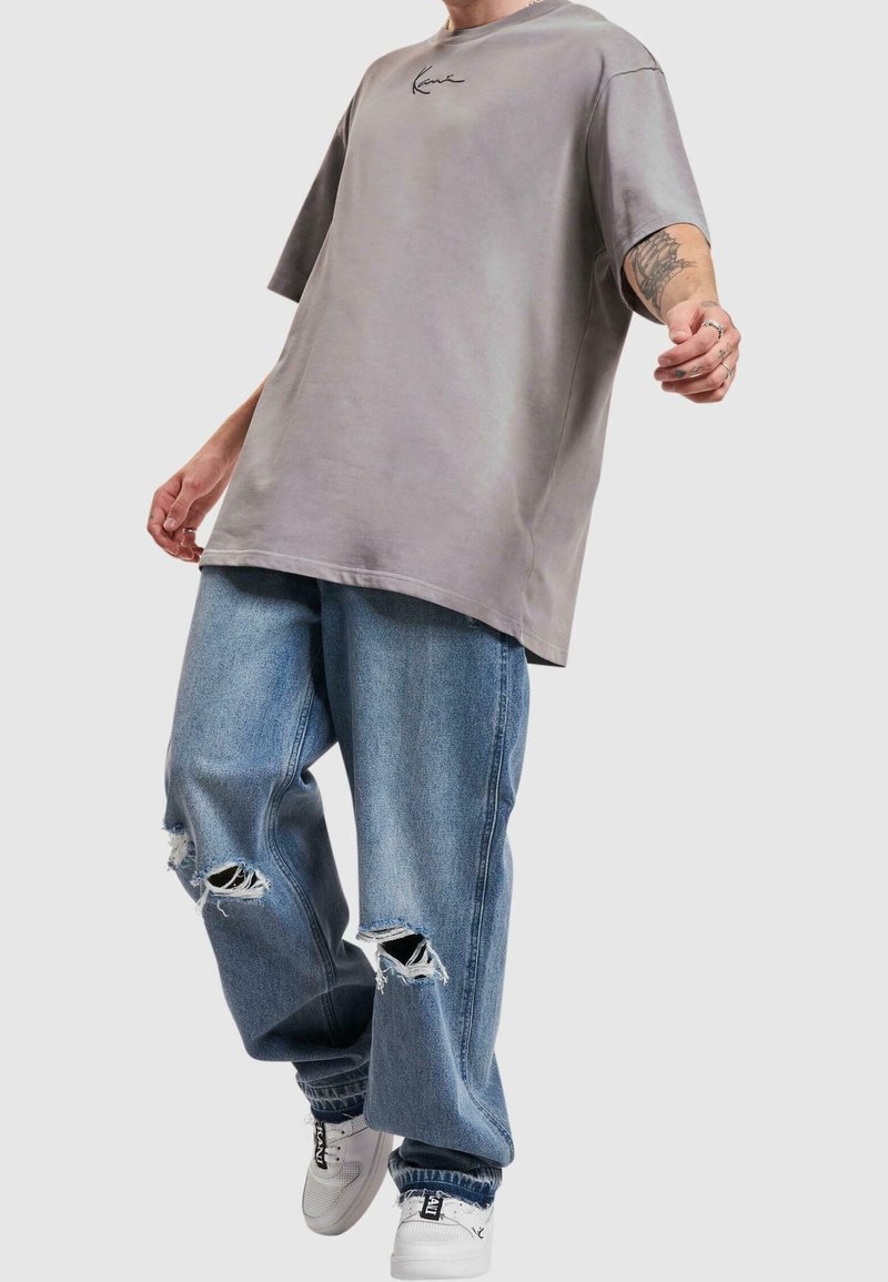 Přes oversized šedé tričko s předním logem, nošené s džíny s lehkým pružením a bílými teniskami. Neformální městský styl.