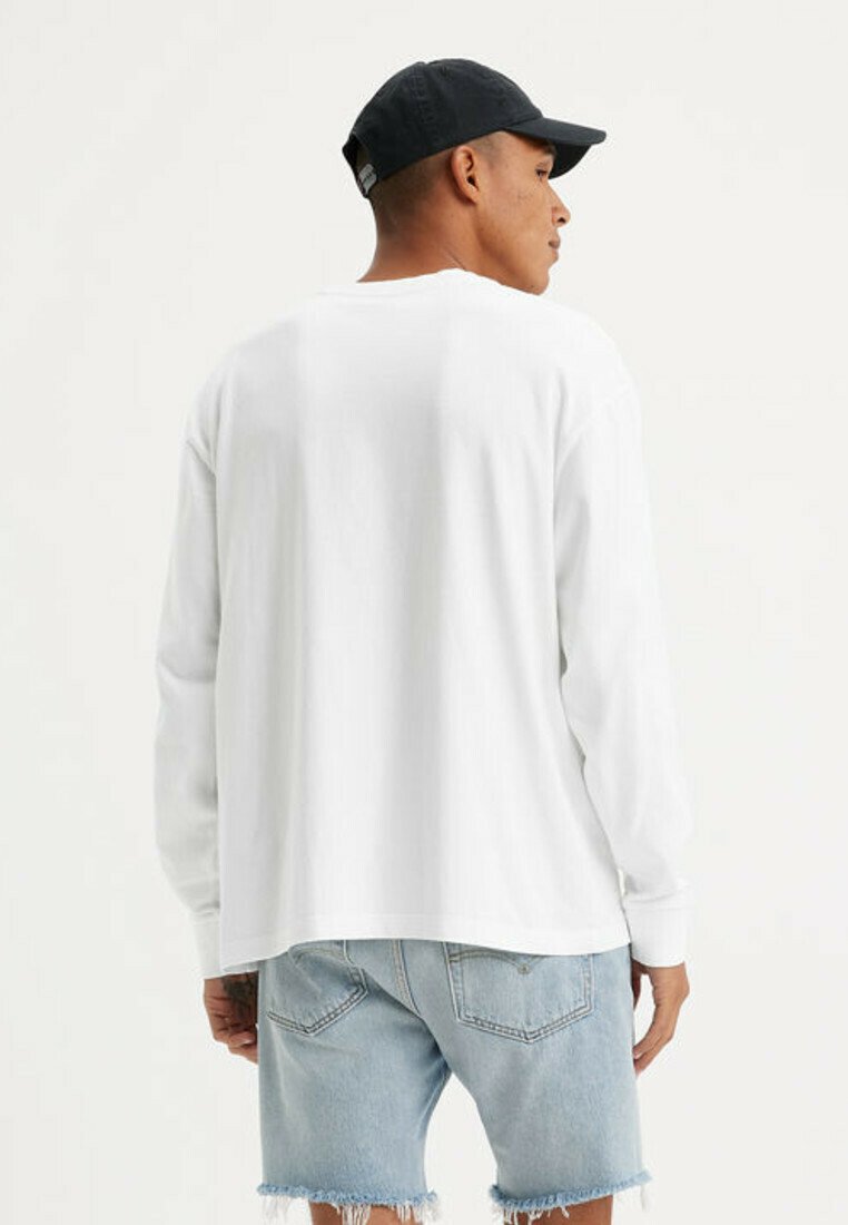Uomo che indossa una camicia bianca a maniche lunghe, pantaloncini di jeans azzurri con bordi sfrangiati e un berretto da baseball nero, visto di spalle.