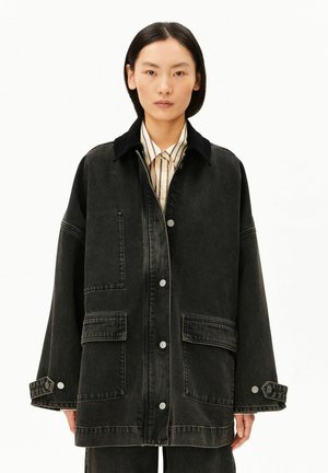 Femme asiatique aux cheveux noirs et raides portant une veste en jean noire oversize sur une chemise beige rayée sur fond blanc.