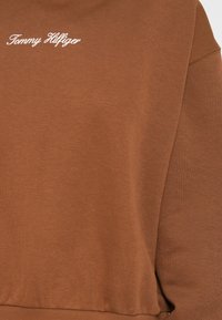 Brun hettegenser med et mykt, teksturert stoff. Har hvit "Tommy Hilfiger" logo brodert på øvre venstre side. Uformell passform.