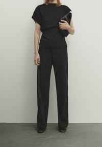 Schwarzes Oberteil mit kurzen Ärmeln und rundem Ausschnitt, kombiniert mit schwarzen, locker sitzenden Hosen. Person hält eine schwarze Clutch. Strukturiertes Gewebe.