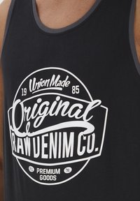 Canotta nera con un logo tondo che presenta "Original RAW DENIM CO." in bianco. Contorno grigio contrastante attorno al colletto. Tessuto morbido.