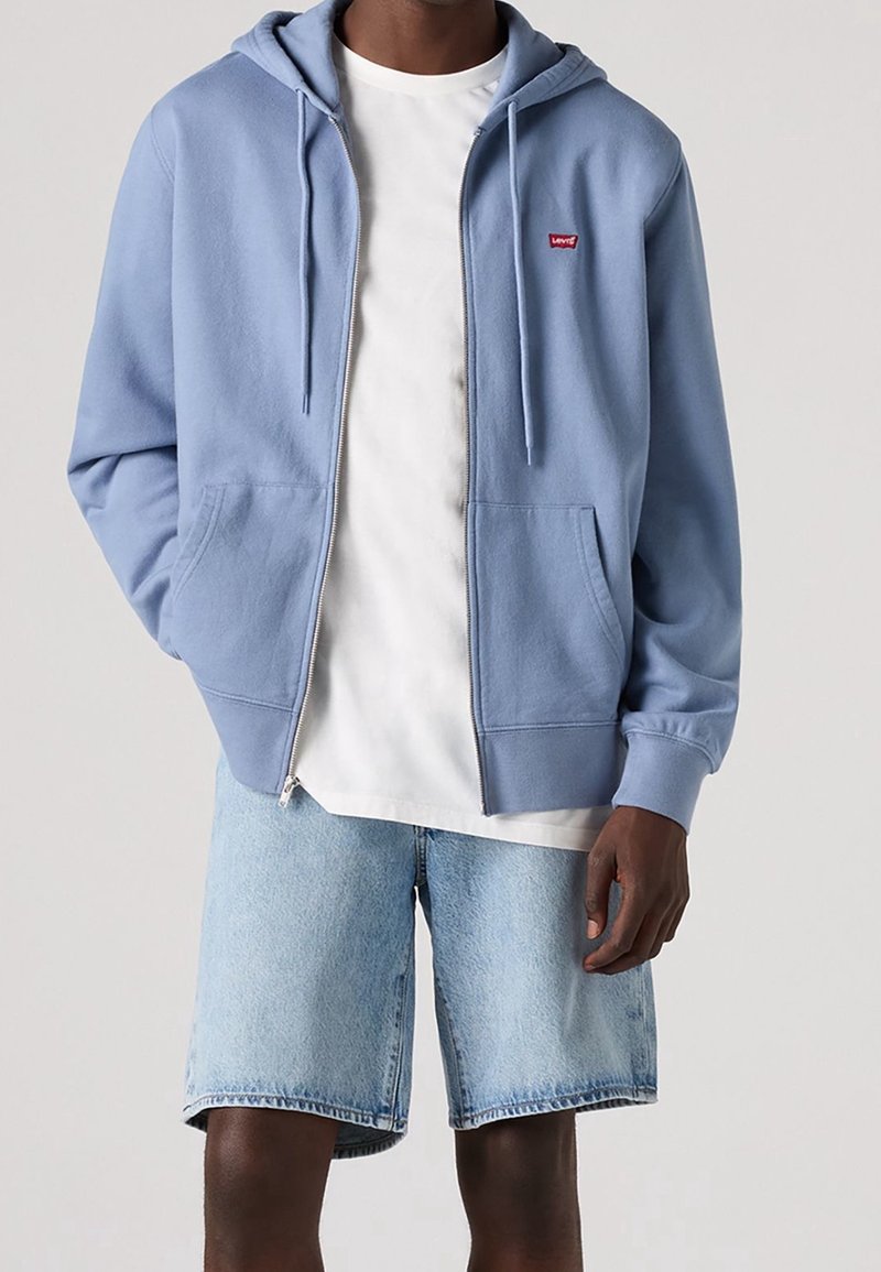 Felpa leggera azzurra con zip, tasca frontale e cappuccio con coulisse, indossata sopra una maglietta bianca, abbinata a pantaloni corti in denim chiaro.