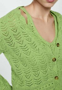 Cardigan vert en tricot avec une texture à motifs de feuilles, col en V et trois boutons marron sur le devant. Conception à épaules ouvertes.