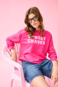 Femme aux cheveux bruns ondulés portant des lunettes noires, un sweat-shirt rose "Sunset Chaser" et un short en jean, assise sur une chaise en plastique rose devant un fond rose.