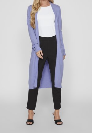 Femme portant un long cardigan lavande, un haut blanc, un pantalon noir court et des sandales noires à talons ouverts, debout devant un fond uni.