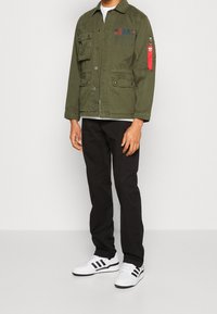 Giacca verde oliva con chiusura a bottoni, multiple tasche e un accento rosso sulla manica. Abbinata a pantaloni neri e sneakers bianche.