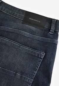 Jean en denim bleu foncé avec une poche arrière et une étiquette en cuir noir indiquant « DENIM SUPPLY Co. » à la taille.