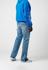 Modrá oversized mikina s bílým textem na rukávu, v kombinaci se světle modrými cargo džínami s několika kapsami a zipovými detaily.