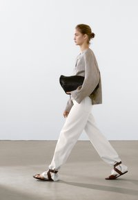 Beige linnen top met brede mouwen, witte ontspannen pasvorm broek, een zwarte geweven clutch in de hand, en bruine sandalen aan. 