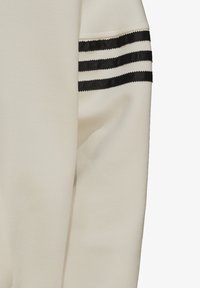 Manica di felpa color crema con una texture liscia, presenta tre righe orizzontali nere e un dettaglio di polsino a coste.