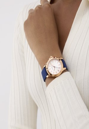 Rose gold watch with a white face, blue silicone strap, and numeric hour markers. Worn on a wrist, with a textured white top in the background.