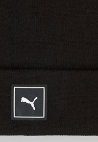 Gorro de punto negro con una etiqueta rectangular negra que presenta un logotipo de gato saltando en blanco. Textura suave y diseño sencillo.