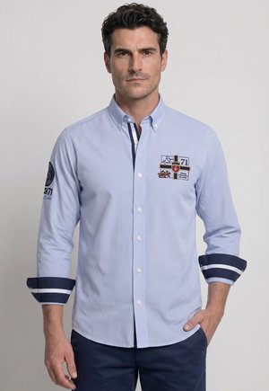 Hombre con camisa de cuadros azul con botones y parches bordados, mangas remangadas que muestran el forro azul oscuro y blanco, de pie con una mano en el bolsillo.