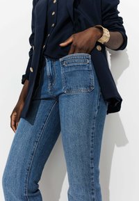 Jean en denim bleu avec une coupe droite, poches avant et arrière, et couture contrastée. Porté avec un blazer bleu marine foncé et un top.
