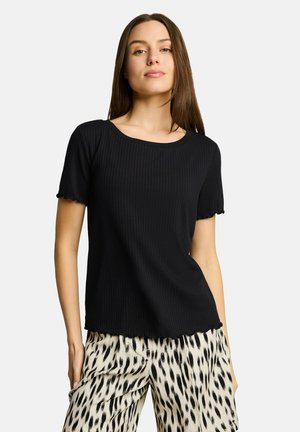 Jeune femme portant un haut noir côtelé à manches courtes et un pantalon blanc ample avec des taches noires abstraites, debout devant un fond blanc.