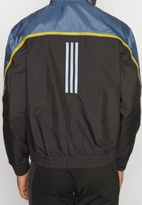 Windbreaker-Jacke mit einem blauen oberen Bereich, einem schwarzen unteren Bereich mit Gitterstruktur, gelben Akzenten und drei hellblauen Streifen auf dem Rücken.