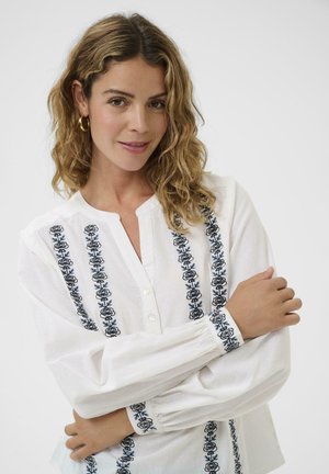 White blouse featuring blue floral embroidery along the front and sleeves, with a buttoned neckline and billowy sleeves. Soft fabric texture.