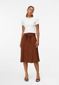 Jupe marron, longueur genou, avec une ceinture à nouer, un bouton à l'avant et des poches latérales, assortie d'une chemise blanche ajustée et de sandales noires.