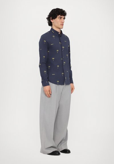 Camicia blu marina con piccolo motivo di palme verdi e la scritta "DREAM", colletto button-down, maniche lunghe, abbinata a pantaloni larghi grigio chiaro.