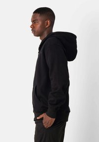 Sudadera negra con capucha, cremallera frontal, puños de canalé y bolsillos; presenta un corte holgado y una textura suave. Vista lateral mostrada.