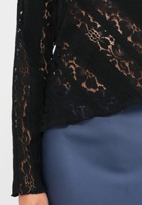 Schwarzes Spitzentop mit komplexen Blumenmustern, strukturiertem Gewebe und einem mehrlagigen Design, kombiniert mit einem glatten, blauen Stoff darunter.