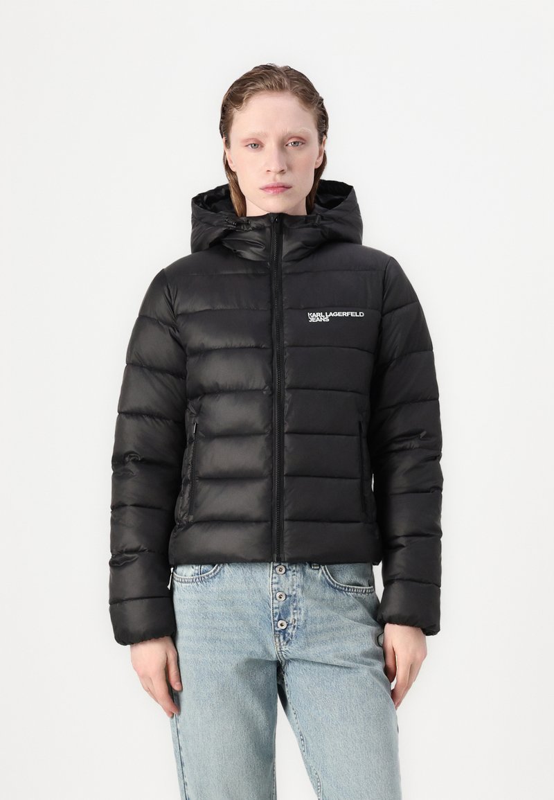 Juodas pūkinis švarkas su gaubtu, siūtas raštu, priekyje užtrauktukas ir šoninės kišenės; ant krūtinės yra logotipas, o kirpimas trumpas.