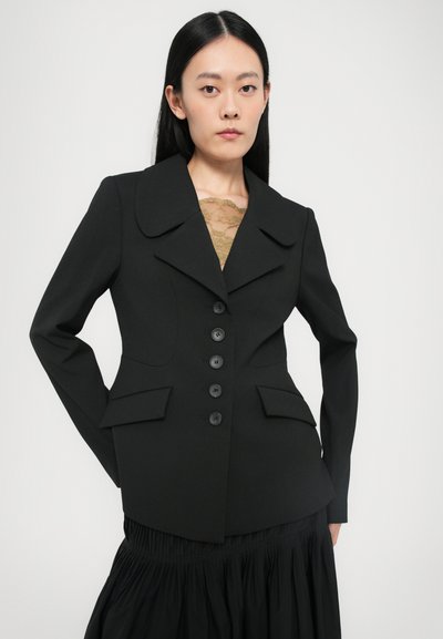Black tailored blazer with large lapels, six black buttons, and two front pockets. Worn over a lace top, paired with a pleated black skirt.