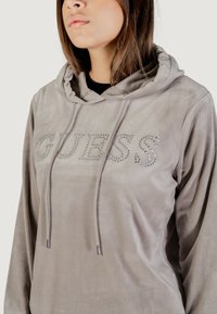 Sudadera gris claro hecha de una tela suave con capucha, que presenta un logo "GUESS" decorado con strass y cordones con puntas metálicas.