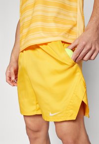 Keltaiset urheilushortsit, jotka on valmistettu kevyestä kankaasta. Niissä on sivutaskut ja pieni valkoinen Nike-logo helman kohdalla. Tennispallo taskussa.