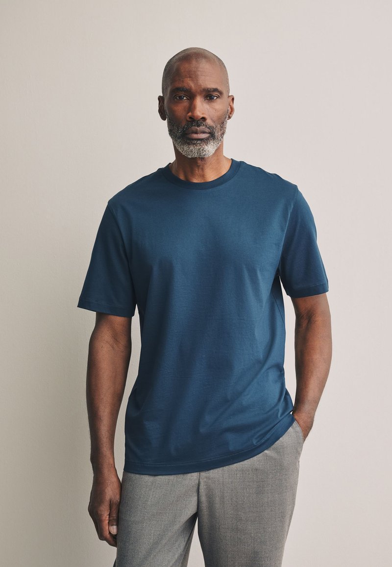Homme avec une barbe grise portant une chemise bleue à manches courtes et un pantalon gris, debout devant un fond clair uni.