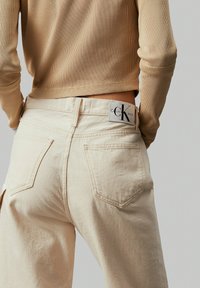 Beež denimpüksid kõrge vöökohaga, viie taskuga ja nahkdetailiga, millel on Calvin Klein Jeans logo. Ribaga beež topp on osaliselt nähtav.