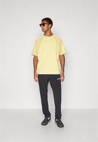 Yellow short-sleeve shirt with contrasting seams, black pants, and dark sneakers. Sunglasses worn, with a simple design and no visible logos.