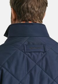 Navy gewatteerde jas met een hoge kraag, voorzien van een zwart GANT-logo patch op de achterkant. Het materiaal lijkt glad en heeft een duurzame afwerking.