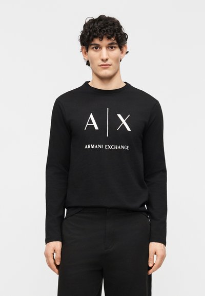 Мъж с къдрави коси, носещ черна дългоръка риза Armani Exchange с бяло лого, стоящ на светъл едноцветен фон.