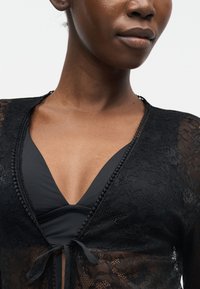 Gros plan d'une personne portant un haut en dentelle noire avec un nœud à l'avant, dévoilant un vêtement intérieur noir en dessous.