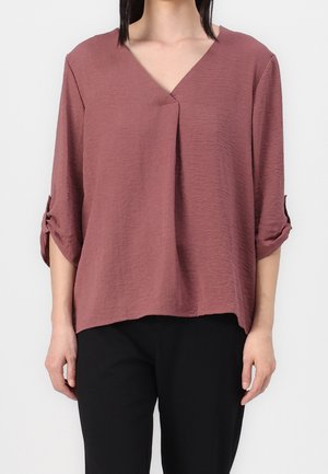 Woman wearing dusty rose V-neck blouse with rolled 3/4 sleeves and black pants, standing against a plain white background.