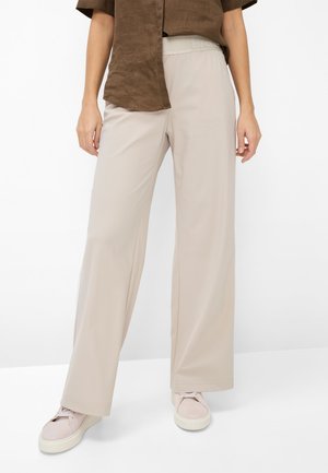 Kvinde iført beige bukser med vidde, brun kortærmet top og lyserøde sneakers, stående foran en ensfarvet hvid baggrund.