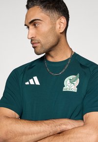 Zöld rövid ujjú sportpóló, mexikói válogatott logóval és fehér Adidas csíkokkal a vállán. Simább felület.