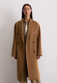 Femme portant un long manteau camel croisé par-dessus une chemise à carreaux marron et un pantalon à carreaux beige, se tenant devant un mur uni.
