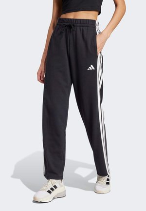 Personne portant un pantalon de survêtement Adidas noir avec des rayures et un logo blancs, associé à des baskets blanches, debout avec une main dans la poche.