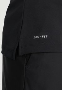 Gros plan sur un t-shirt de sport noir avec "DRI-FIT" imprimé en blanc près de l’ourlet, superposé à un pantalon noir.