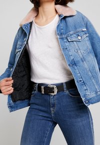 Giacca di jeans blu chiaro con colletto rosa pallido, tasche frontali e fodera trapuntata nera, indossata sopra una t-shirt bianca e jeans blu.