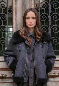 Giacca in denim nera con collo e polsini in pelliccia sintetica, chiusura a zip e tasche frontali, abbinata a una camicia nera a righe.