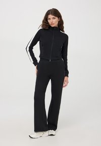 Czarna kurtka zapinana na zamek z wysokim kołnierzem i dwoma przednimi kieszeniami, zestawiona z czarnymi spodniami dzwonami. Białe paski boczne akcentujące rękawy.