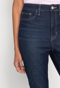 Los jeans de mezclilla azul oscuro presentan un diseño de cintura alta con bolsillos frontales y costuras amarillas en contraste. Textura suave con un ligero estiramiento.