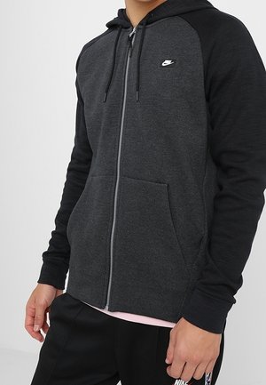 Mand iført mørkegrå og sort Nike hættetrøje med lynlås, forlommer og et lille hvidt Nike-logo på brystet.