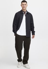 Svart skjorte med knapper over en hvit T-skjorte, kombinert med mørke bukser og hvite sneakers. Et brunt mønstret skjerf er båret rundt halsen.
